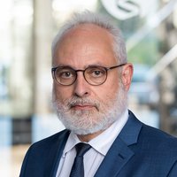 Der Arzt Dr. Siegfried Krell trägt einen dunkelblauen Anzug, weißes Hemd und schwarze Krawatte, steht vor einem modernen Gebäude