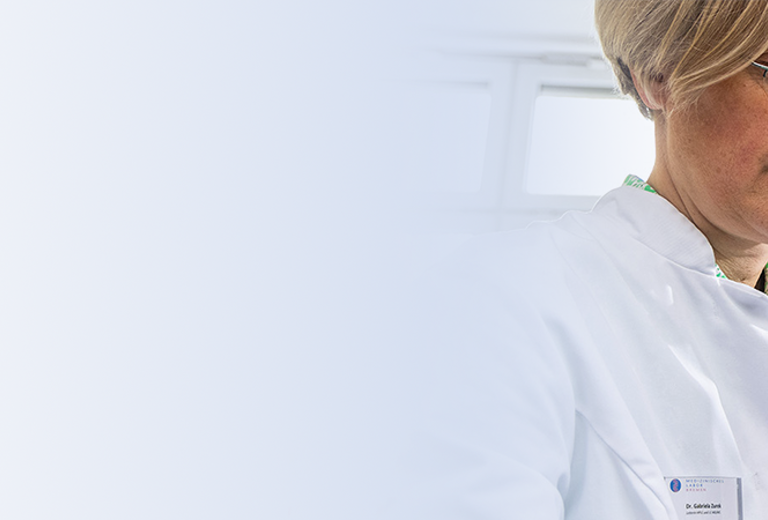 Zwei Laborantinnen in weißen Laborkitteln, eine hält ein Tablet, die andere steht im Hintergrund. Im Hintergrund sind technische Geräte sichtbar.