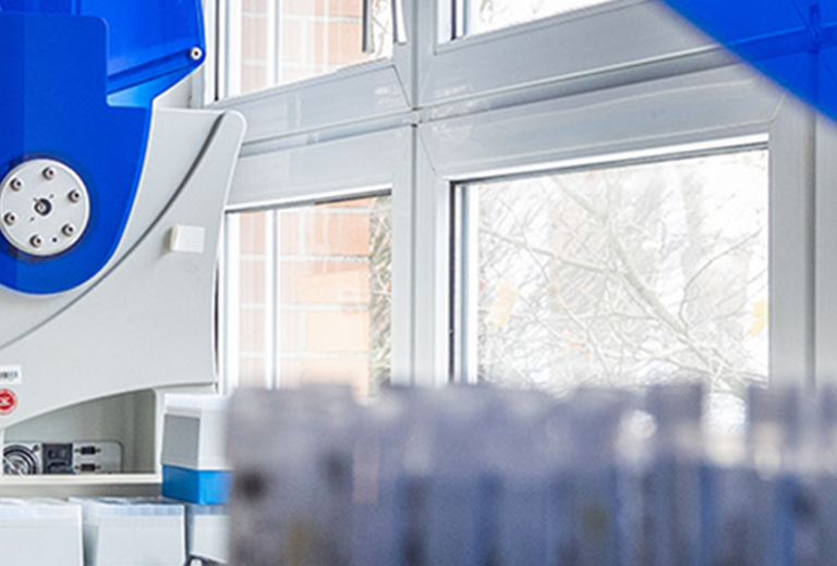 Laborumgebung mit einer Labortechnikerin, die an einem großen, modernen Analysegerät arbeitet. Im Vordergrund sind Reagenzgläser sichtbar.