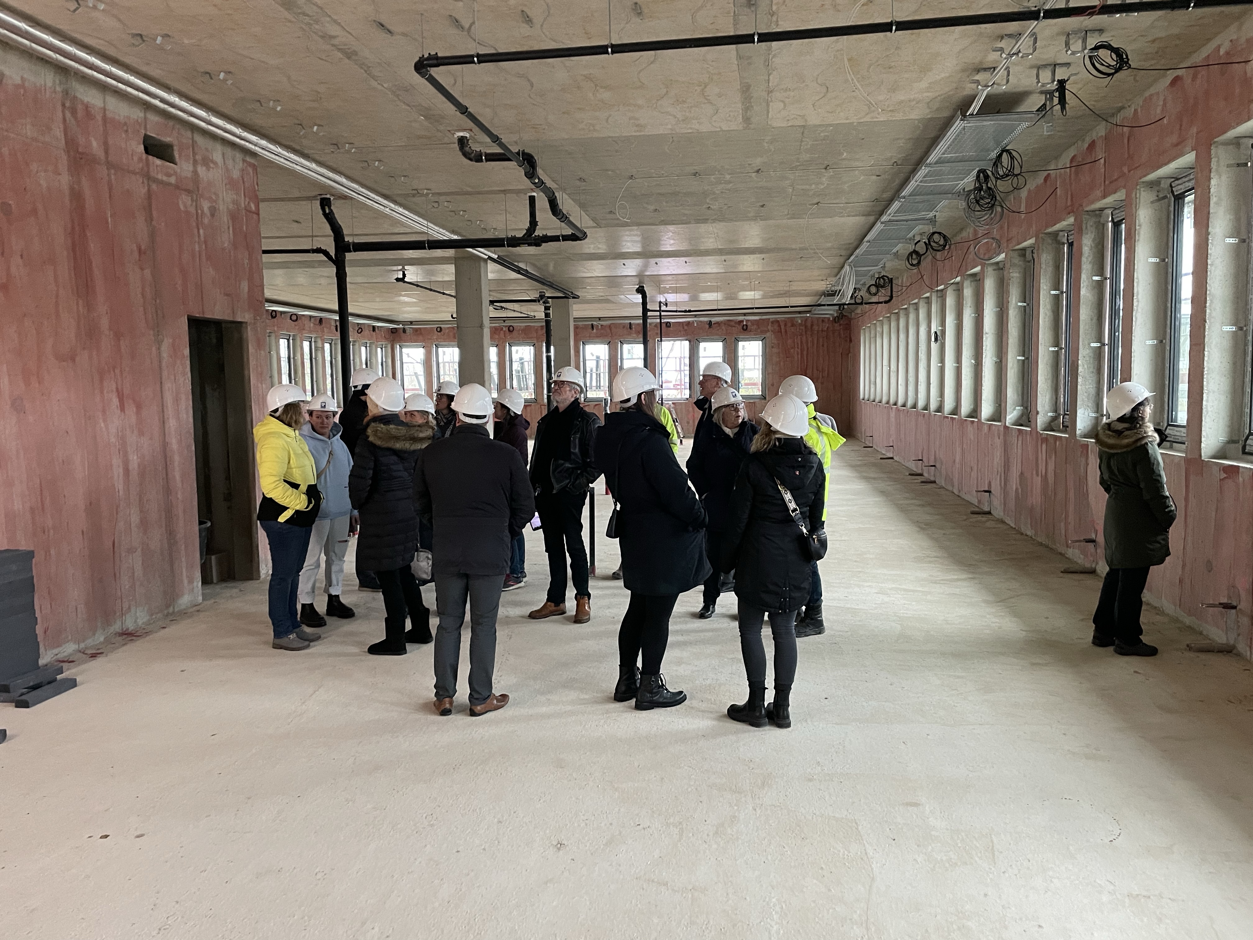 Gruppe von etwa 15 Personen mit weißen Schutzhelmen steht in Rohbau mit freiliegenden Wänden und Decke, großen Fenstern und sichtbaren Rohrleitungen.