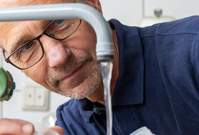 Mann mit Brille füllt im Rahmen mikrobiologischer Wasseruntersuchungen eine Probe aus einem Wasserhahn in ein klares Gefäß