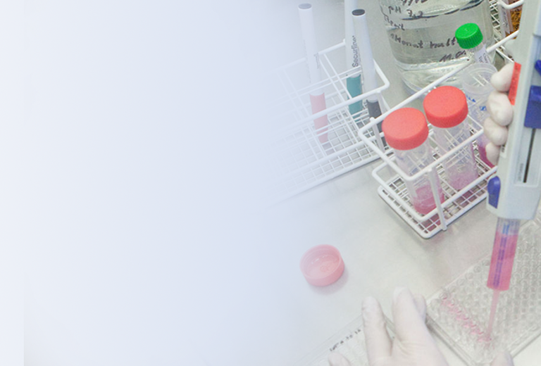 Eine Laborantin in einem weißen Laborkittel führt einen Serologietest durch, indem sie eine Pipette in ein Reagenzglas mit lila Flüssigkeit eintaucht. Im Hintergrund sind mehrere Reagenzgläser und ein Labortisch sichtbar.