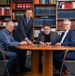 Vier formell gekleidete Mitarbeiter sitzen an einem runden Holztisch vor einem Bücherregal und blicken in die Kamera