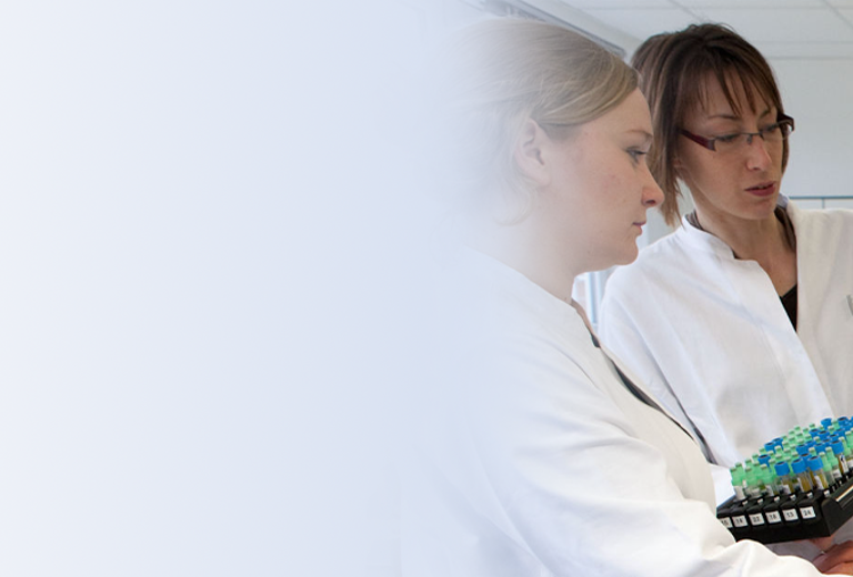 Zwei Laborantinnen in Laborkitteln stehen neben einem Computer und halten ein Probenrack mit mehreren Röhrchen in der Hand.