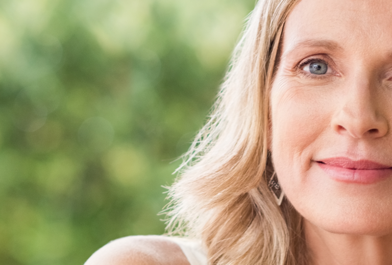 Eine ältere Frau mit langen blonden Haaren sitzt entspannt auf einer Bank im Freien, trägt ärmelloses weißes Oberteil