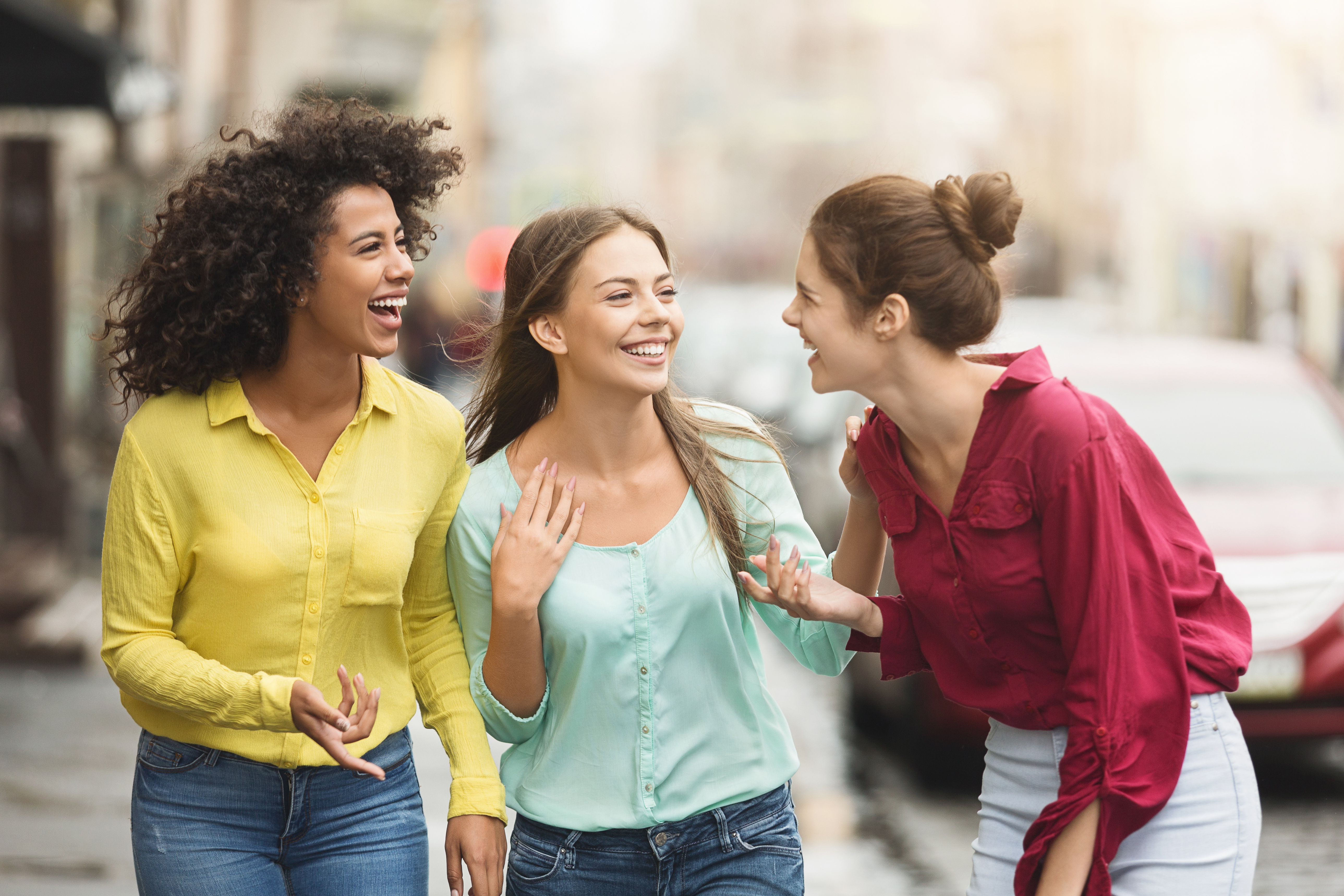 Drei Frauen gehen auf einer Straße, zwei tragen bunte Blusen in Gelb und Pink, eine trägt eine grüne Bluse, sie unterhalten sich lebhaft