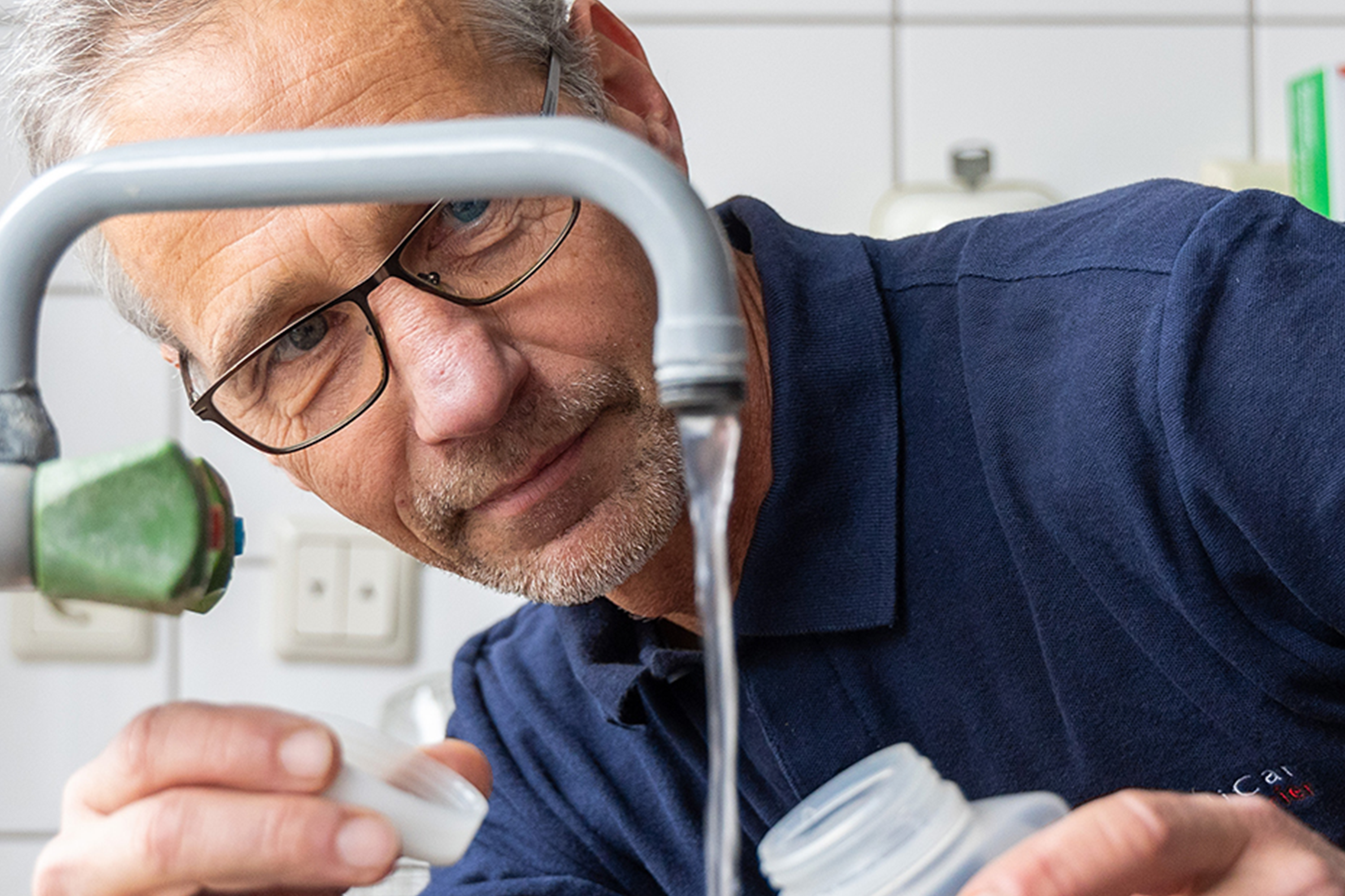 Mann mit Brille füllt im Rahmen mikrobiologischer Wasseruntersuchungen eine Probe aus einem Wasserhahn in ein klares Gefäß ab