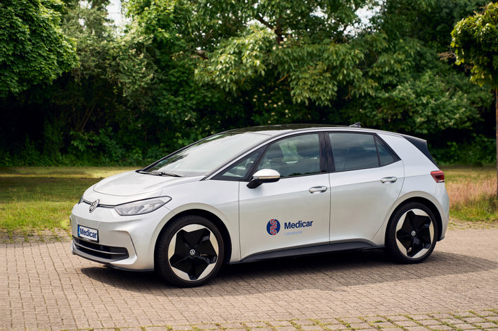 Ein silbernes Auto steht auf einem gepflasterten Weg. Auf der Seite ist das Logo 'Medicar' sichtbar. Im Hintergrund sind Bäume zu sehen.